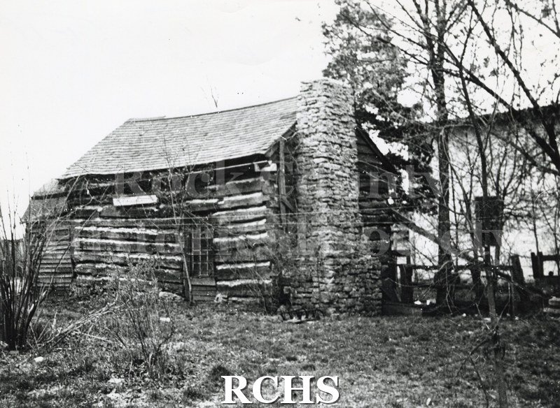 Black and white photograph of Pioneer log cabin. Cabin has one door and a brick chimney on the outside of the house on the right side.