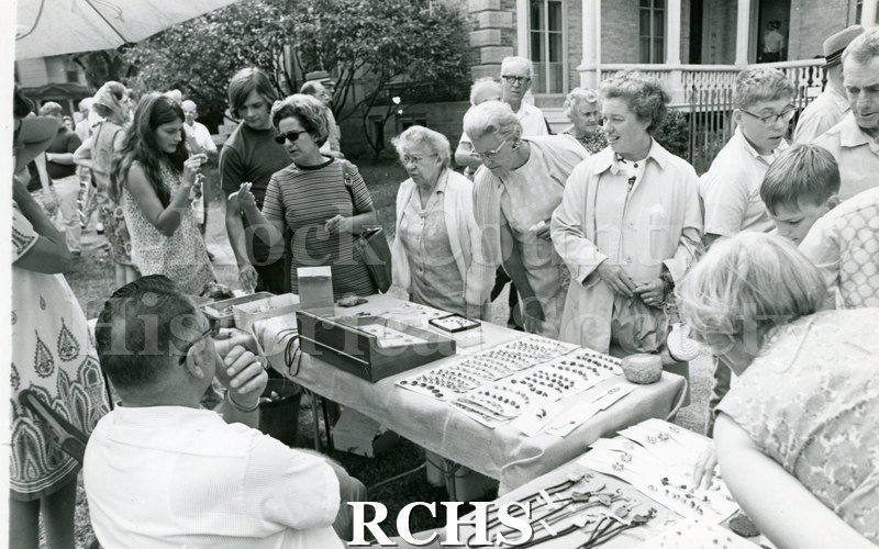Black and white photograph of the 14th Annual Ice Cream Social & 1st Arts & Crafts Fair. People are standing around a booth that is selling jewelry.