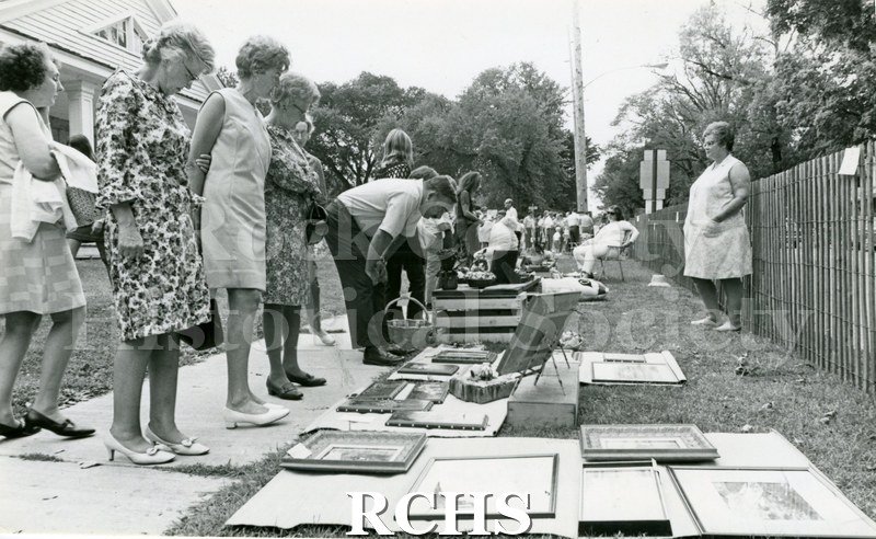 Black and white photograph of the 14th Annual Ice Cream Social & 1st Arts & Crafts Fair". People are standing and looking at paintings displayed on the ground.