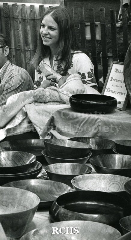 Black and white photograph of the 1972 Arts & Crafts Fair. Shows a mother feeding her baby a bottle. She is seated in a booth with wooden bowls in front.