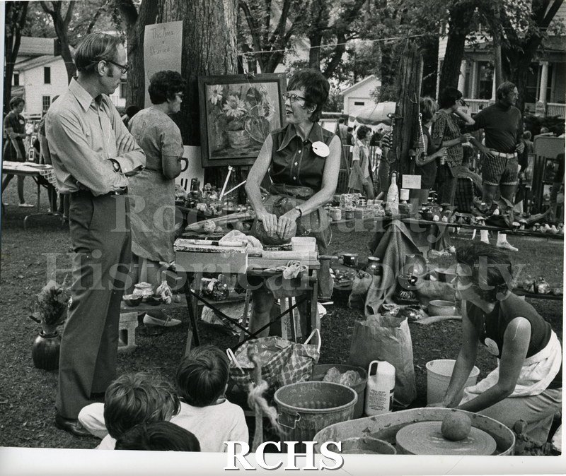 Black and white photograph of the 1972 Arts & Crafts Fair. Shows a woman talking with a man. She appears to be demonstrating some skill.