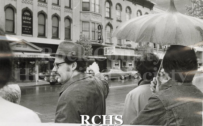 Black and white photograph of walking tour of downtown Janesville with Richard Hartung and others. Across the street is "IOOF" and "Hall's". Mr.
