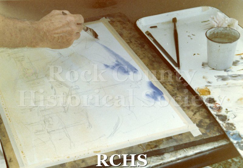 Color photo of Don Johanning painting. Only the hand with the brush is visible. There is a cup of water and two paint brushes on a tray to the right.