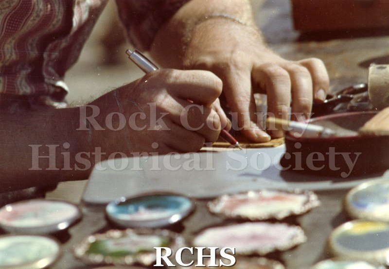 Color photo of Don Johanning writing. Only two hands are visible. Other items are on the table as well as a cigarette in an ashtray.