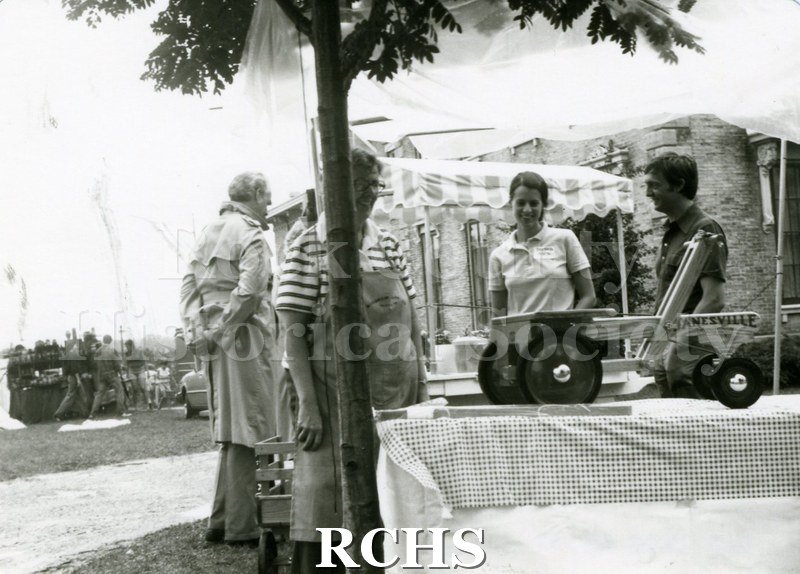 Black and white photograph of several people standing around an exhibit of a Janesville Wagon which is sitting on a table.
