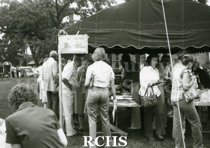 Black and white photograph of several people standing around a tent in which ice cream and cake are sold.