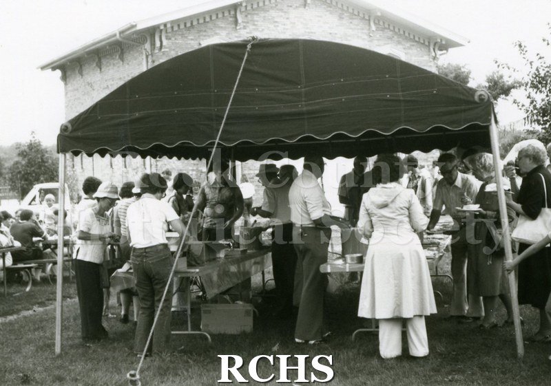 Black and white photograph of several people standing around a tent in which food is being sold.
