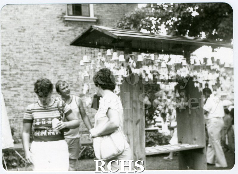 Black and white photograph of people standing around a booth selling bells.