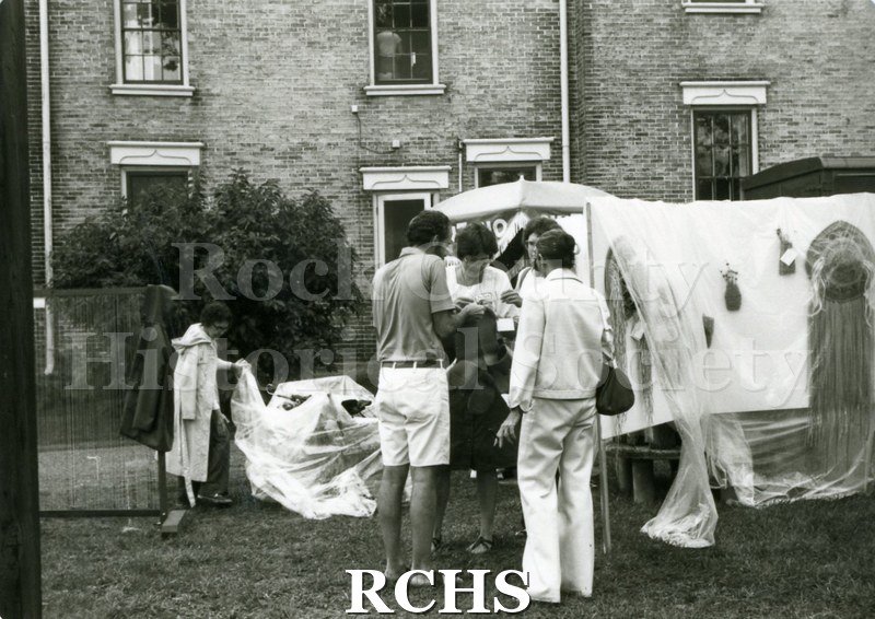 Black and white photograph of people standing around a booth draped in plastic to shield the artifacts for sale from the rain..
