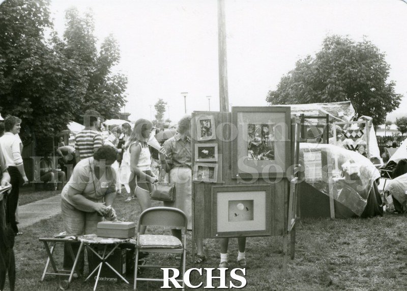 Black and white photograph of people getting ready for the festival. Several paintings are displayed in the middle of the photograph.