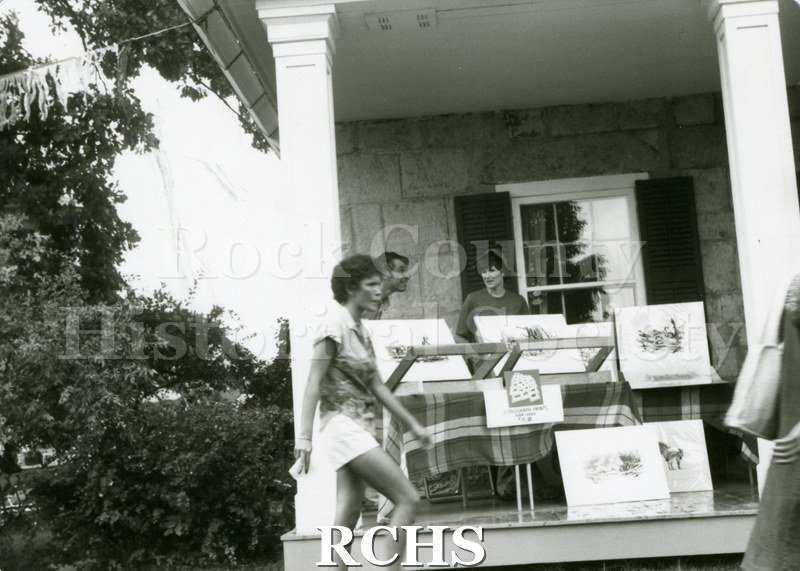 Black and white photograph of people standing near an exhibit of paintings on the porch of the stone house at the Tallman Arts Exhibit.