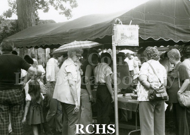 Black and white photograph of people standing around tables selling food at the Tallman Arts Exhibit.