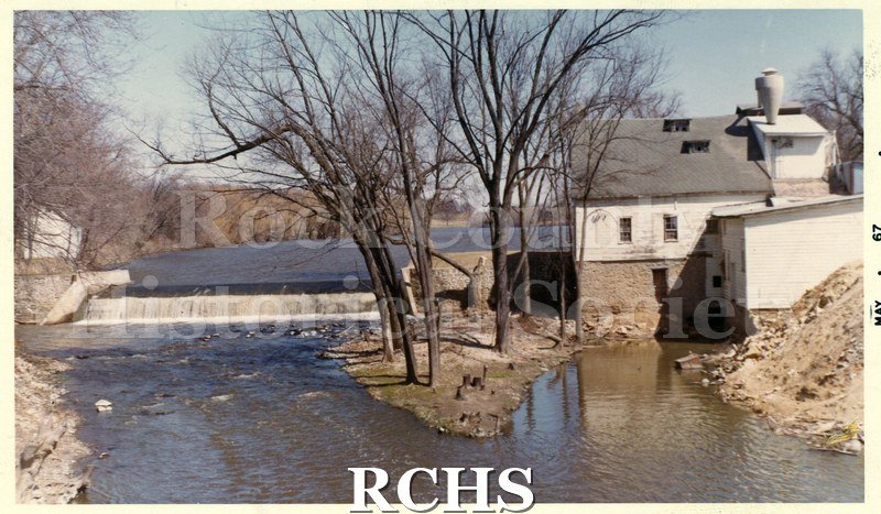 Colored photograph of Rockdale Mill, Dane Co. There is a dam on the left side of the photograph and a building on the right.