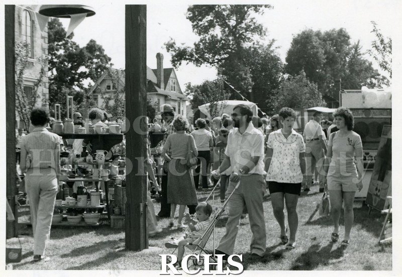 Black and white photograph of the 1981 Tallman Festival. People are looking at an exhibit of pottery. A man in the foreground has a baby in a stroller.