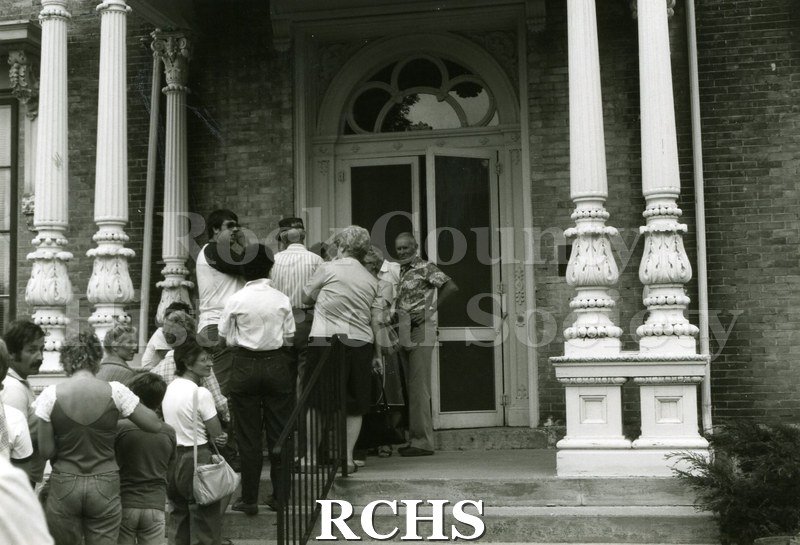 Black and white photograph of the 1983 Tallman Festival. Shows people on the porch of the Tallman House. N83013 Strip.