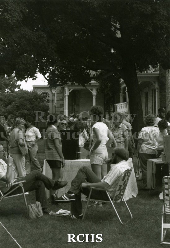 Black and white photograph of the 1983 Tallman Festival. Shows people sitting and standing around the Tallman House exhibits. N83013 Strip.