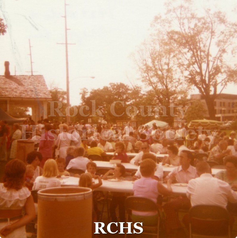 Colored photograph of people sitting at tables and others standing at the 1977 Art Festival.