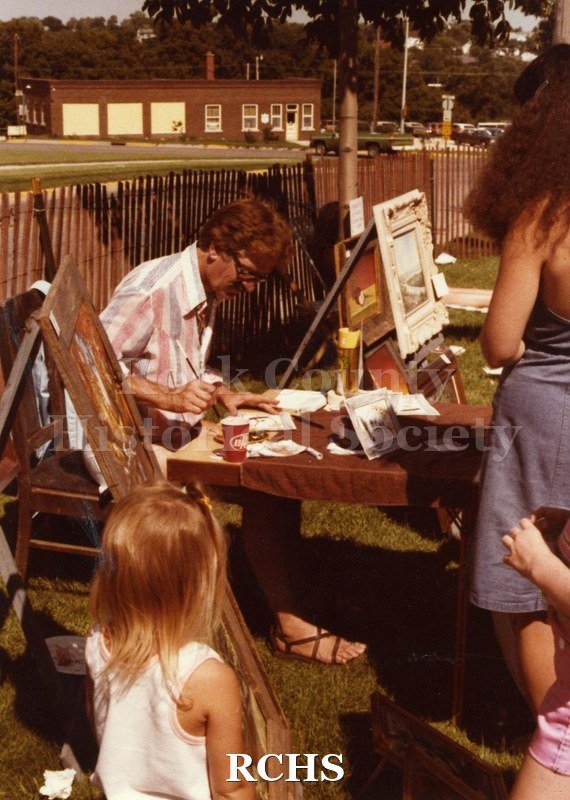 Colored photograph of an unidentified woman working on an art project while people look on at the Art Festival.