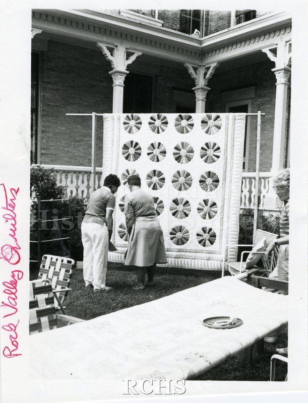 Black and white photograph of two unidentified women examining a quilt displayed on the lawn near the Tallman House. On side: "Rock Valley Quilters".