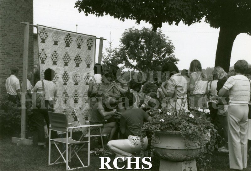 Black and white photograph of people looking at a quilt while others are waiting.