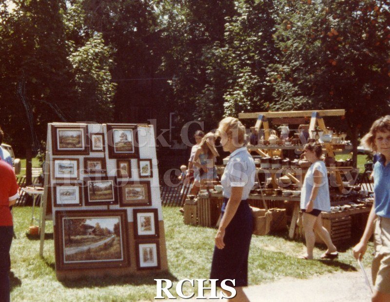 Colored photograph of people looking at a display of paintings. A display of pottery is on the right.
