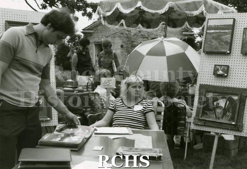 Black and white photograph of an unidentified man looking through a notebook while the artist sits at a table.There are other exhibiters in the background.