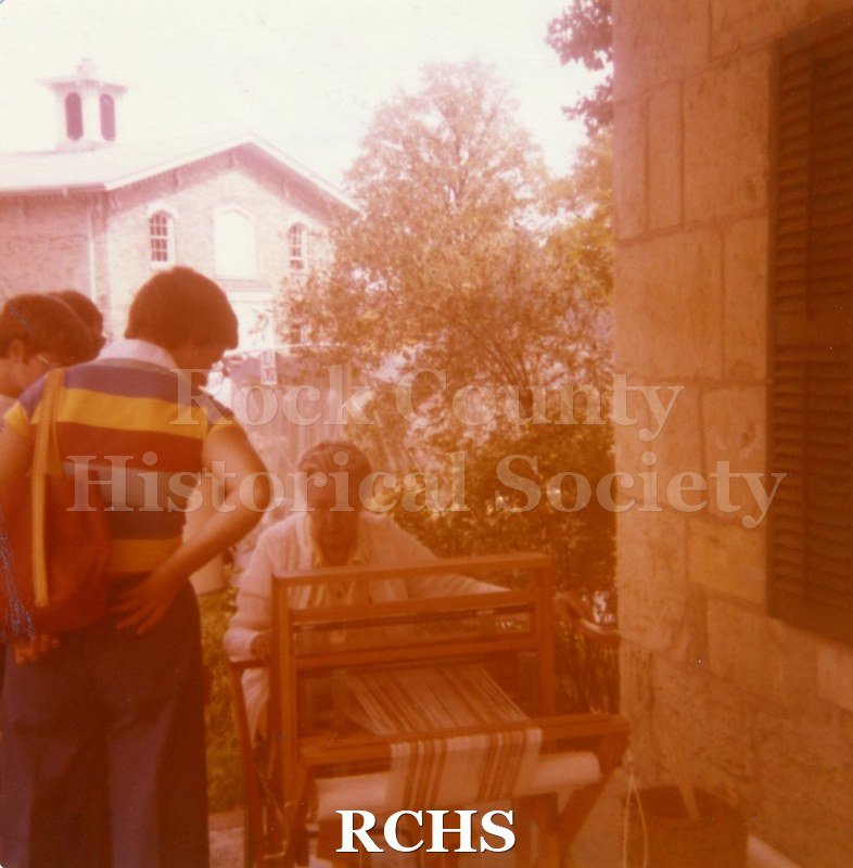 Colored photograph of the 1977 Arts & Craft Fair. Shows people watching a woman at a loom.