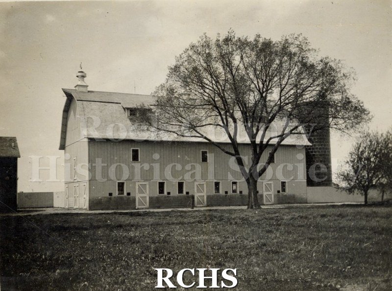 Black and white photograph of an unidentified two story barn with a silo. Barn has windows on both floors. A tree is directly in front of the barn.