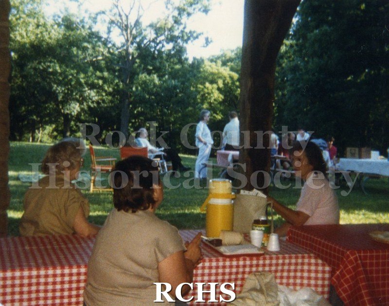 Color photo of Rock County Historical Society Picnic. Three women are seated at a table and other people are in the background.