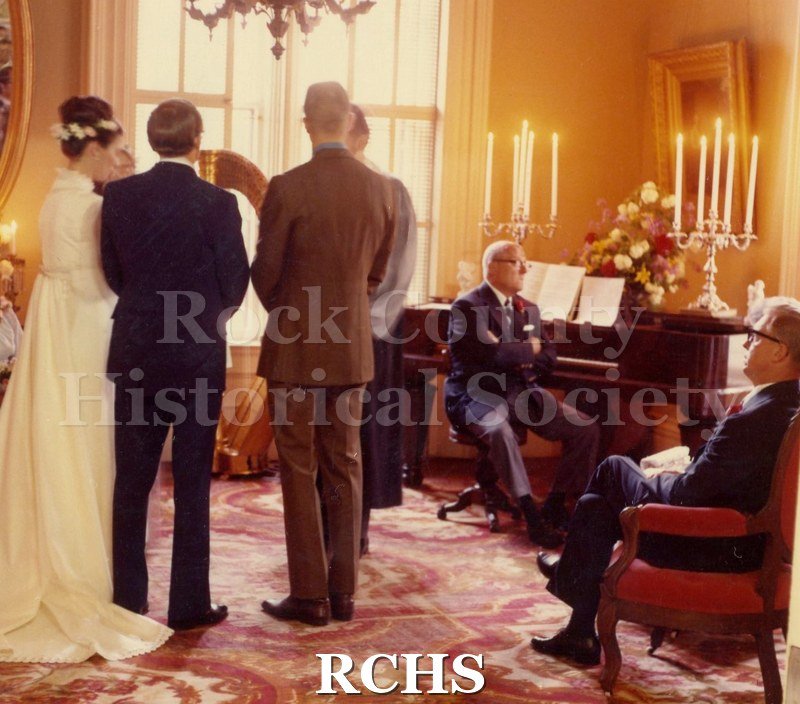 Color photo of bride and groom standing in the Tallman House. There is a man sitting at the piano and another is sitting in a chair.