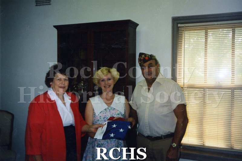 Color photo of two women and a man. The person in the middle is holding a folded flag.