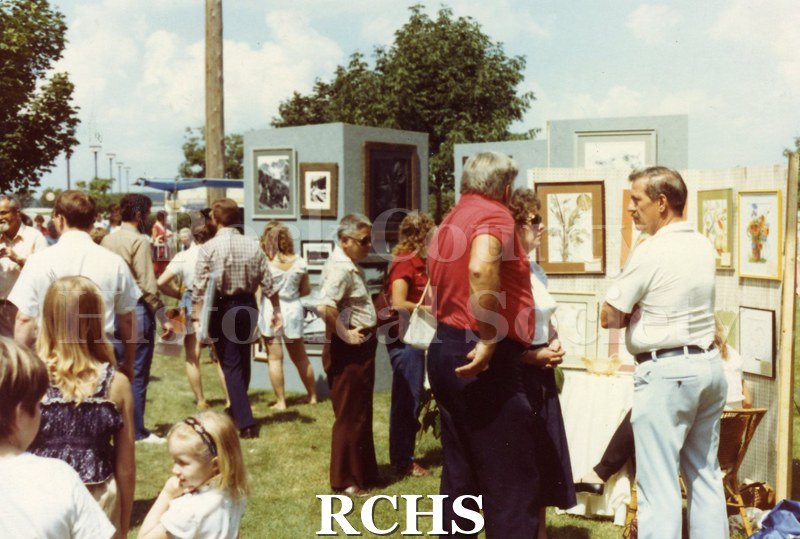 Colored photograph of people strolling the grounds at the Tallman Arts Festival. Exhibits of paintings are in the background.