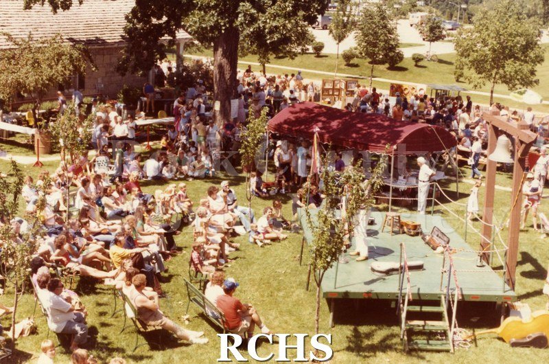 Colored photograph of the 1982 Tallman Arts Festival, taken from above. Shows stage on the right and people sitting on chairs in front of it.