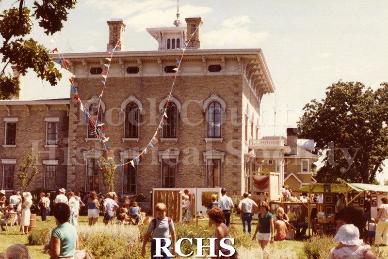 Colored photograph of the Tallman Arts Festival taken at ground level. The Tallman House is visible in the background with pennants draped from the chimneys.