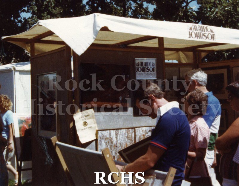 Colored photograph of an unidentified man and woman standing near a display booth. The canvas on top reads: "Alaine Johnson".