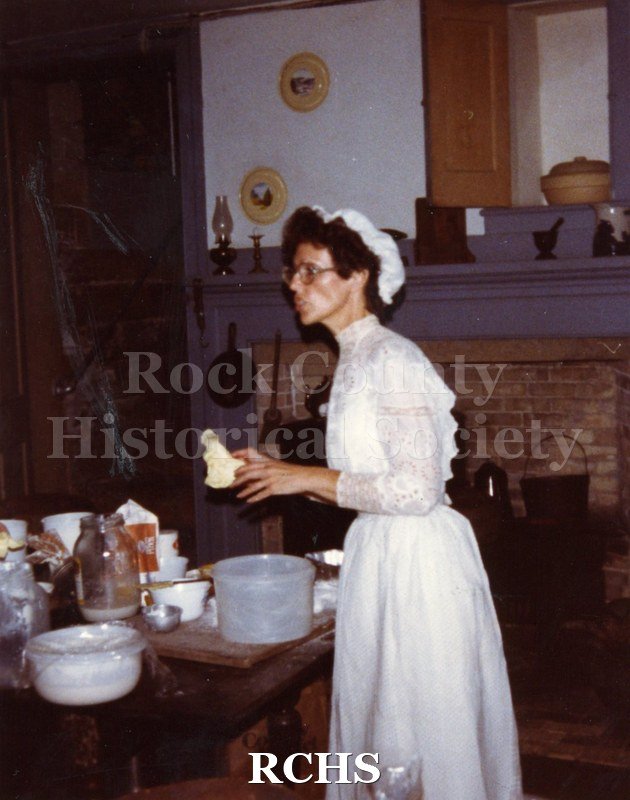 Colored photograph of June Loper wearing a period costume making bread in the basement kichen.