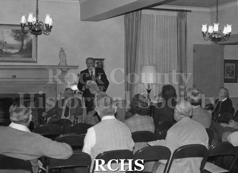 Black and white photograph of several people sitting in a room in the Tallman House. Kenneth Bick is giving a talk.