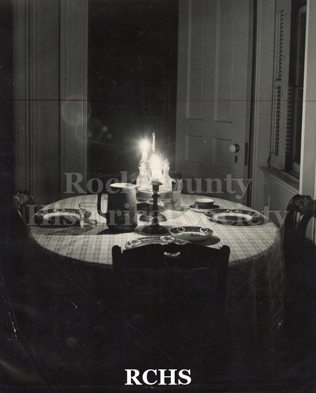 Black and white photograph of a table with candles aglow on it. Table is set with china. The rest of the room is dark.