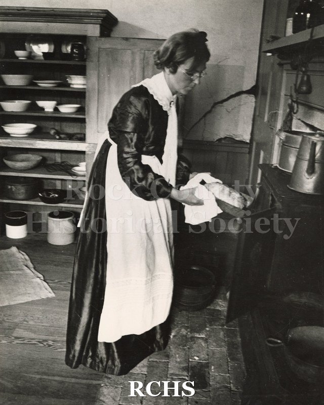 Black and white photograph of June Loper dressed in period costume taking a loaf of bread out of the oven. There is a cupboard displaying bowls behind her.