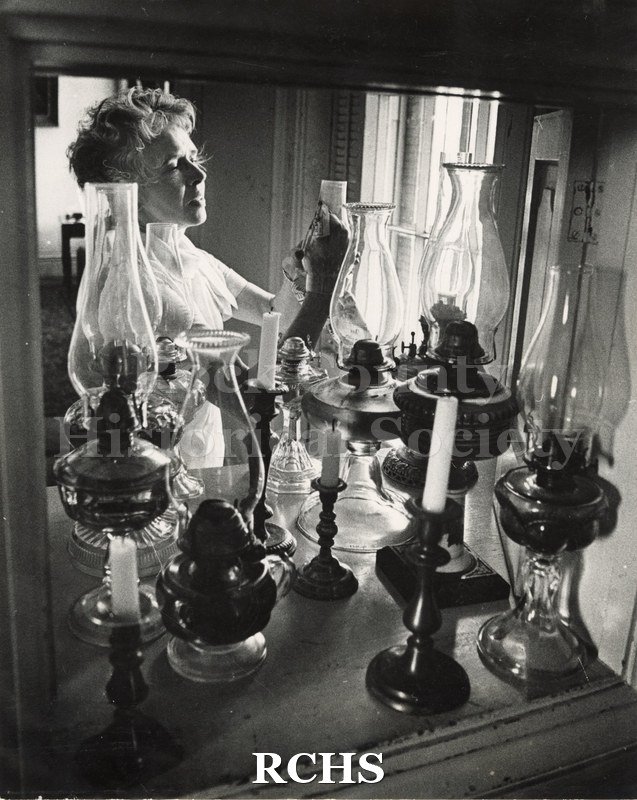 Black and white photograph of Ruth Hughes in period dress lighting a lantern. There are many lanterns and several candles on the table in front of her.