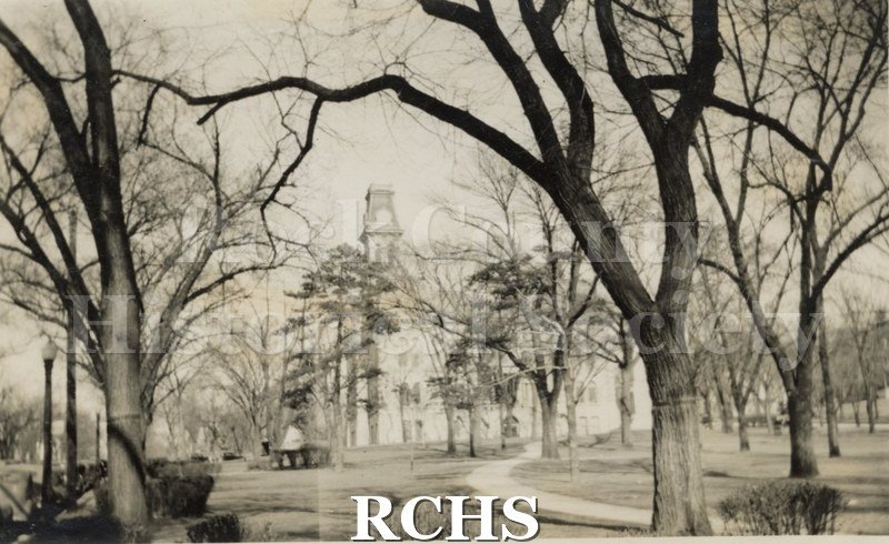 Black and white photograph of the Rock County Court House with trees in front of it and a curved sidewalk. There is a man sitting on a bench on the left side.