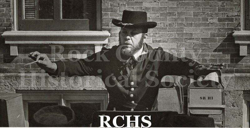 Black and white photograph of a band director wearing a Civil War era uniform. He has his arms out directing.