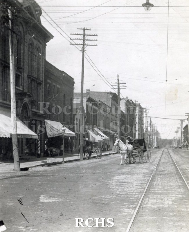 Black and white photograph of Main Street. Shows a horse and carriage being driven along a street with trolley tracks in the middle and buildings on the sides.