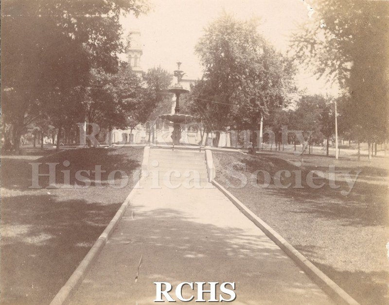 Black and white photograph of the Rock County Court House, Janesville, WI. Shows Court House Park and the sidewalk leading up to the fountain.
