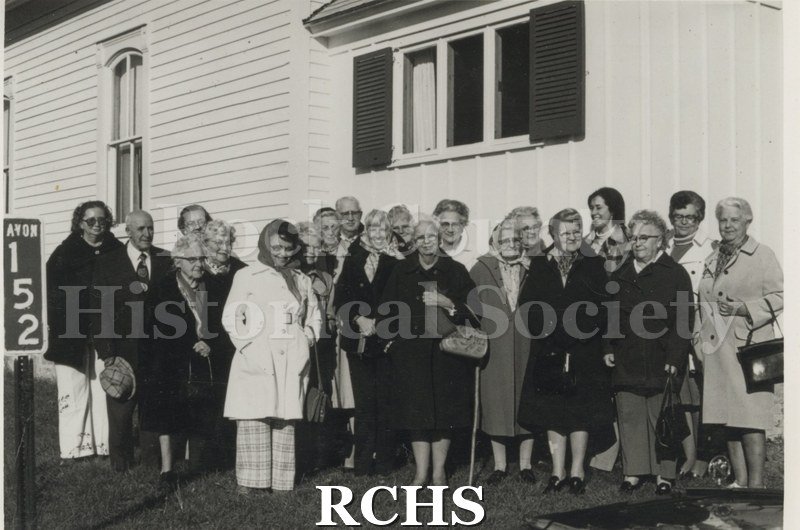 Black and white photograph of the Rock County Historians, Avon Church. Shows several women and men standing beside a building. They are wearing winter clothes.