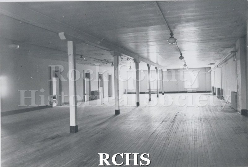 Black and white photograph of Myers Block-Old Armory, Janesville, WI, interior view, showing pillars holding up a beam. There are several doors on the left side.