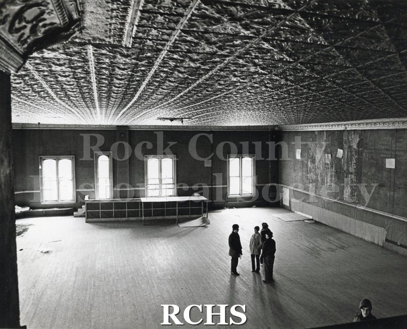 Black and white photograph of Myers Block-Old Armory, Janesville, WI, interior view with five people inside. Shows the ceiling and arched windows.
