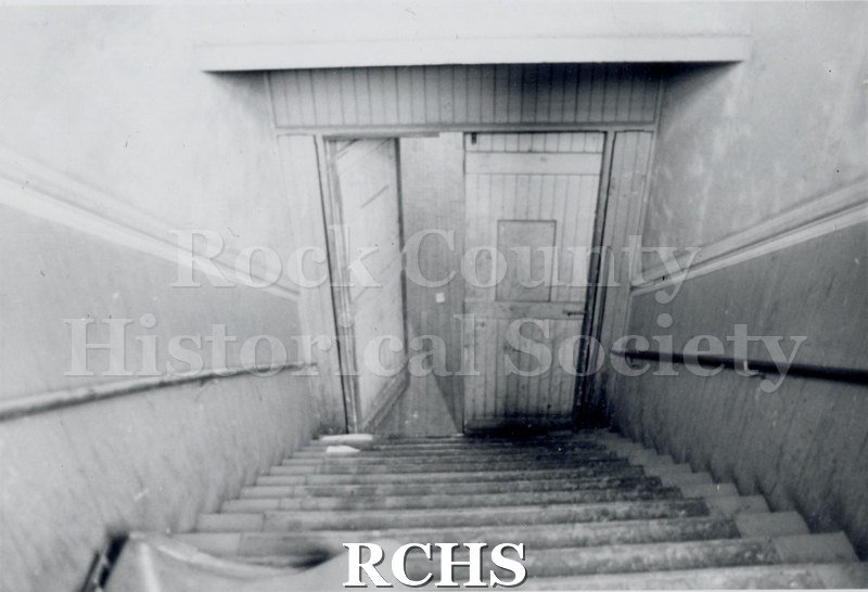 Black and white photograph of Myers Block-Old Armory, Janesville, WI, interior view. There are stairs going down to a doorway.