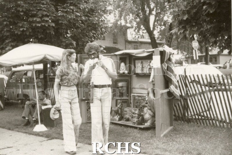 Black and white photograph of the Tallman Arts Exhibit. Shows two people on the sidewalk in front of the exhibits. The man is eating.
