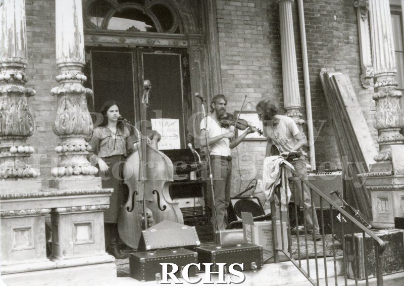 Black and white photograph of the Tallman Arts Exhibit. On the Tallman House porch are three people in a band.